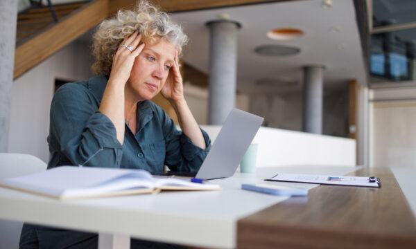 Professional woman suffering workplace stress, pressing fingers to temples while working on laptop in contemporary corporate setting