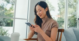 A young Asian woman is sitting in a cafe, using her smartphone to bank online.