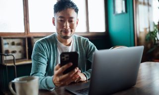 Confident young Asian man looking at smartphone while working on laptop computer in home office.