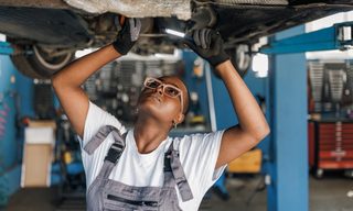 A young African American female mechanic is standing under a car and examining the damage on the broken parts with a serious look on her face.